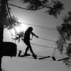 Silueta de una persona cruzando un puente colgante de madera en un circuito de arvorismo a gran altura, con el sol de fondo y rodeado de árboles en Santo Amaro. Silhueta de uma pessoa atravessando uma ponte pênsil de madeira em um circuito de arvorismo alto, com o sol ao fundo e rodeada por árvores em Santo Amaro.