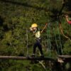 Niño con casco amarillo cruzando un obstáculo de troncos en el circuito de arvorismo, supervisado de cerca por una instructora en una plataforma de madera en medio de la selva. Criança com capacete amarelo atravessando obstáculo de troncos no arvorismo, supervisionada de perto por uma instrutora em plataforma de madeira na mata.