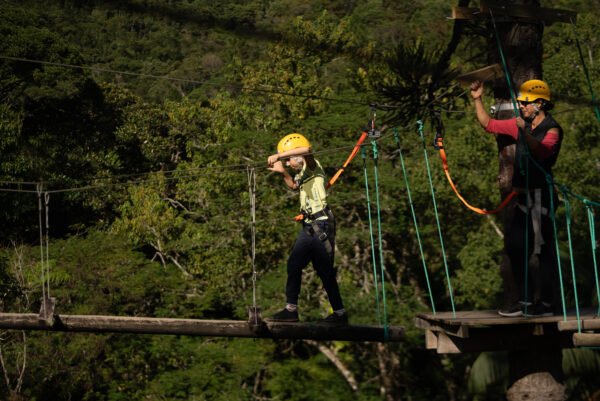 Niño con casco amarillo cruzando un obstáculo de troncos en el circuito de arvorismo, supervisado de cerca por una instructora en una plataforma de madera en medio de la selva. Criança com capacete amarelo atravessando obstáculo de troncos no arvorismo, supervisionada de perto por uma instrutora em plataforma de madeira na mata.
