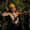 Primer plano de una mujer sonriendo y disfrutando, equipada con casco y arnés, agarrándose de las cuerdas de seguridad en el circuito de arvorismo rodeada de vegetación. Sinta a liberdade de se movimentar em meio à Mata Atlântica com total segurança e muitas risadas. Close de uma mulher sorrindo e se divertindo, equipada com capacete e cadeirinha, segurando nas cordas de segurança do circuito de arvorismo em meio à vegetação.