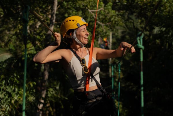 Primer plano de una mujer sonriendo y disfrutando, equipada con casco y arnés, agarrándose de las cuerdas de seguridad en el circuito de arvorismo rodeada de vegetación. Sinta a liberdade de se movimentar em meio à Mata Atlântica com total segurança e muitas risadas. Close de uma mulher sorrindo e se divertindo, equipada com capacete e cadeirinha, segurando nas cordas de segurança do circuito de arvorismo em meio à vegetação.