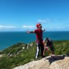 Turista haciendo rapel con los brazos abiertos mostrando seguridad en el equipo mientras el instructor ajusta el arnés, con vista panorámica al mar y morros de Florianópolis.