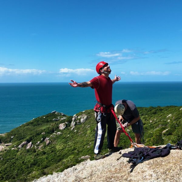 Turista haciendo rapel con los brazos abiertos mostrando seguridad en el equipo mientras el instructor ajusta el arnés, con vista panorámica al mar y morros de Florianópolis.