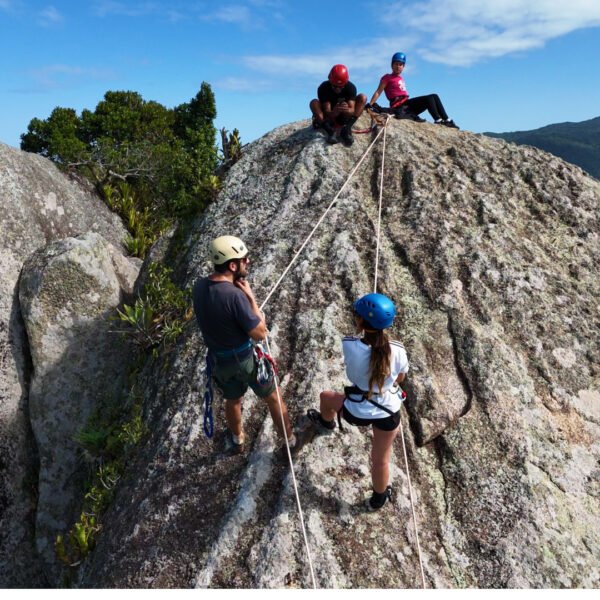Instructor supervisando a un turista realizando el descenso de rapel en una roca alta de Praia Mole, Florianópolis, con grupo esperando y equipo de seguridad completo.